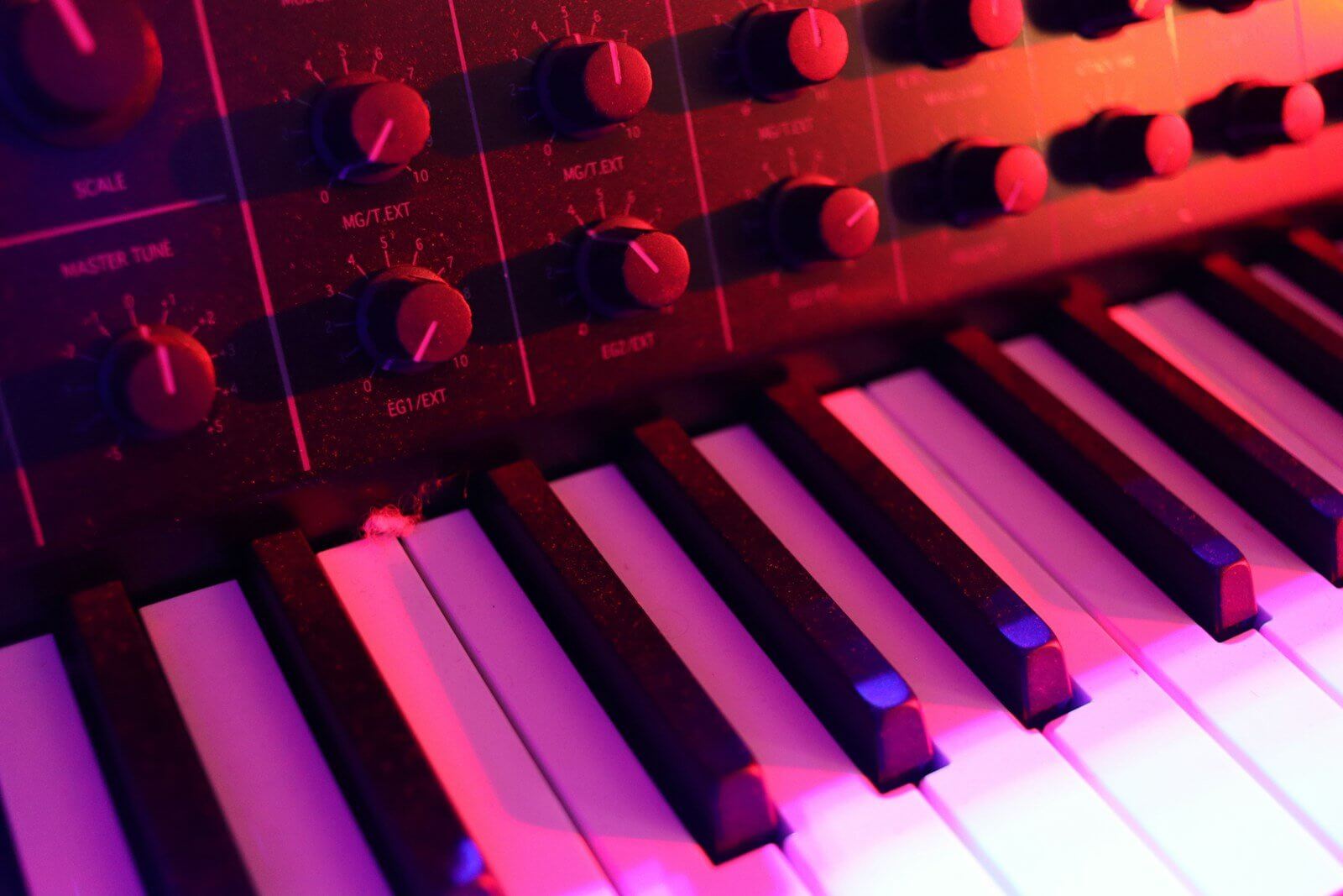 Close-up of an analog synthesizer, showing its knobs and keys under red and purple light